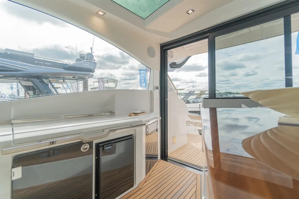 Interior view of a yacht featuring a modern kitchenette with a fridge, large windows, and a wooden deck outside.